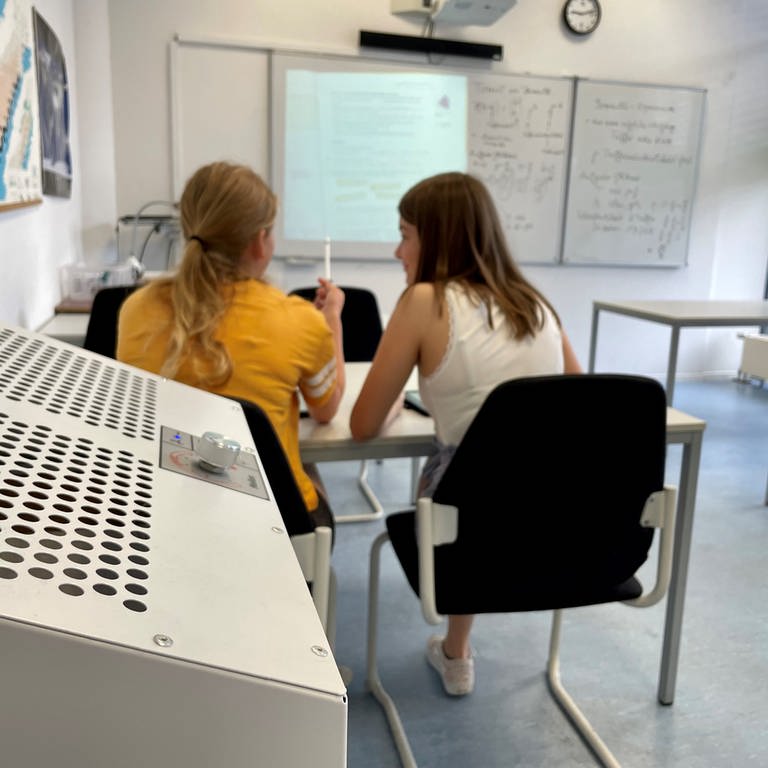 Ein mobiler Luftfilter steht in einem Klassenraum, im Hintergrund sitzen zwei Schülerinnen. (Foto: SWR, Wera Engelhardt)