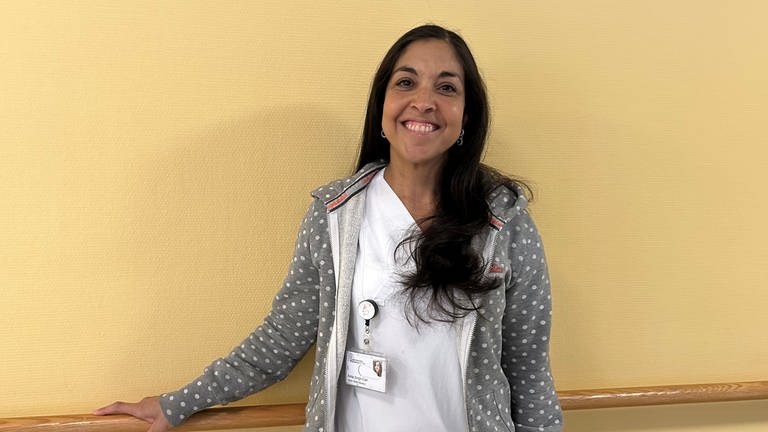 Anita Singh-Carl steht in einem weißen Krankenpflegerinnen-Outfit vor einer gelben Wand in der Uniklinik.