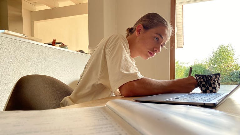 Linus trägt ein weißes T-Shirt und sitzt an einem Esstisch. Vor ihm liegen sein Laptop und sein Mathebuch. Auf dem Laptop ist die Nachhilfe-App "Matheheld" zu sehen. Linus schaut konzentriert auf den Laptop und hält einen Stift in der Hand. Er hat seine blonden Haare zu einem Zopf gebunden. (Foto: SWR, Katharina Seeburger)