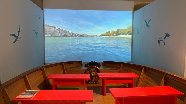 Im Vordergrund stehen vier rote Sitzbänke je zwei neben einander. Die Sitzbänke stehen in einem Boot. Vor dem Boot hängt eine Leinwand. Darauf ist eine Filmaufnahme von einer Schifffahrt auf dem Rhein bei Basel zu sehen.  (Foto: SWR, Katharina Seeburger)