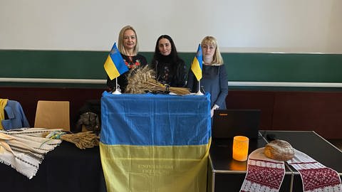 Drei Frauen stehen nebeneinander. Vor ihnen steht ein Stehpult. Darüber ist die blau-gelbe Fahne der Ukraine gehängt. Auf dem Tisch liegt ein Bünde getrockneter Ähren. (Foto: SWR, Sebastian Bargon)