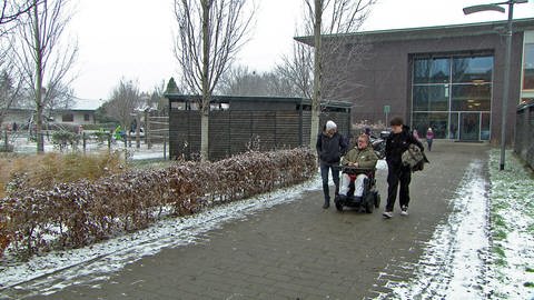 Junge mit blonden Haaren und Brille sitzt in einem selbstfahrenden Rollstuhl. Neben ihm ein Mann in Jacke.  (Foto: SWR)