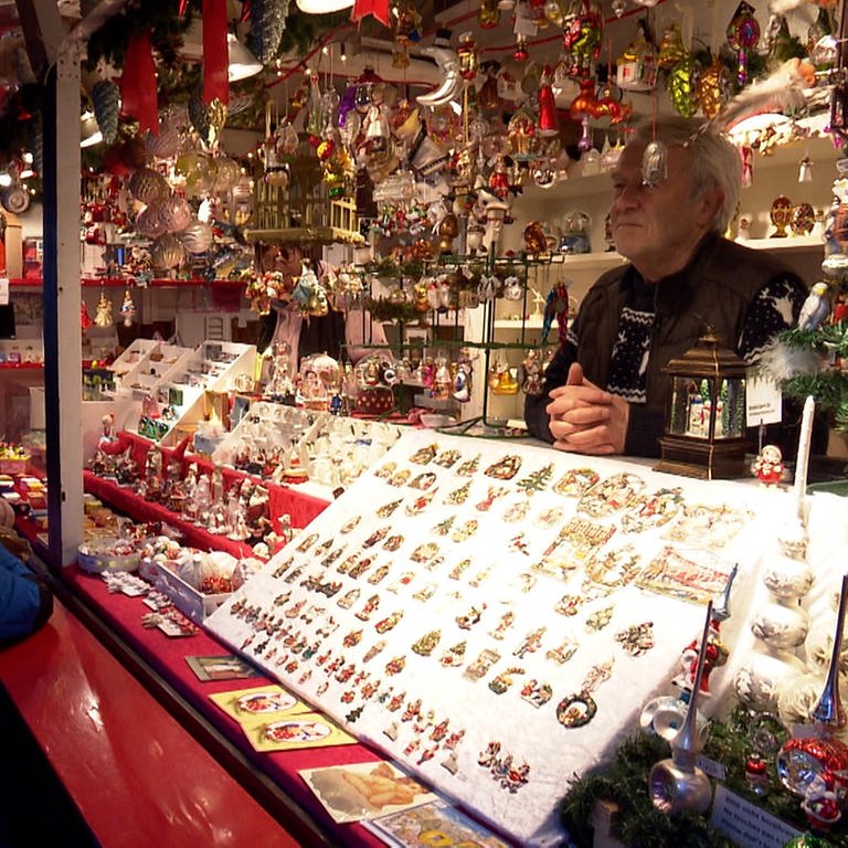 In einer Weihnachtsmarkt Bude hängen viele bunte Kugeln. Im inneren steht ein Mann mit wießen Jahren. 