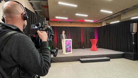 Martin Horn steht hinter einem Rednerpult auf einer Bühne. Er trägt einen schwarzen Anzug. Links im Bild ist ein Kameramann zu sehen. Er filmt Martin Horn. (Foto: SWR, Nadia El-Gonemy)
