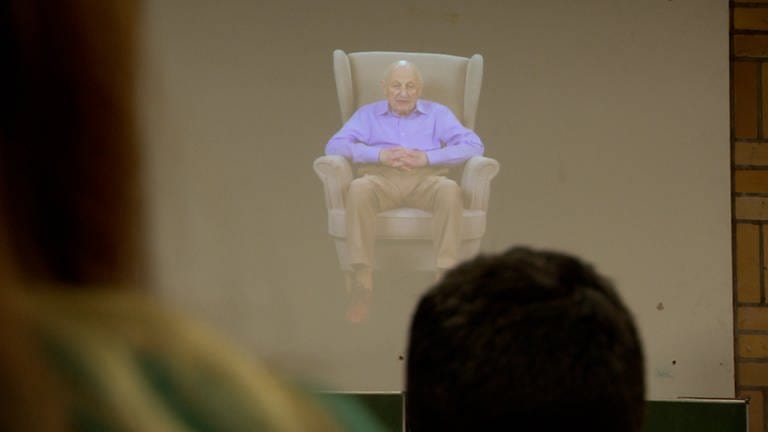Ein älterer Mann in einem lila Hemd sitzt auf einem großen Sessel.