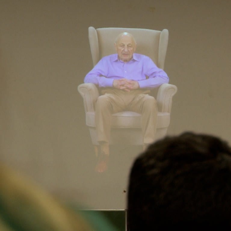 Ein älterer Mann in einem lila Hemd sitzt auf einem großen Sessel.