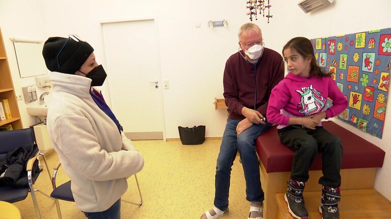 Eine Frau mit Maske ganz links steht in einem Behandlungszimmer, auf der Liege sitzt ein Mädchen, dahinter der Arzt.