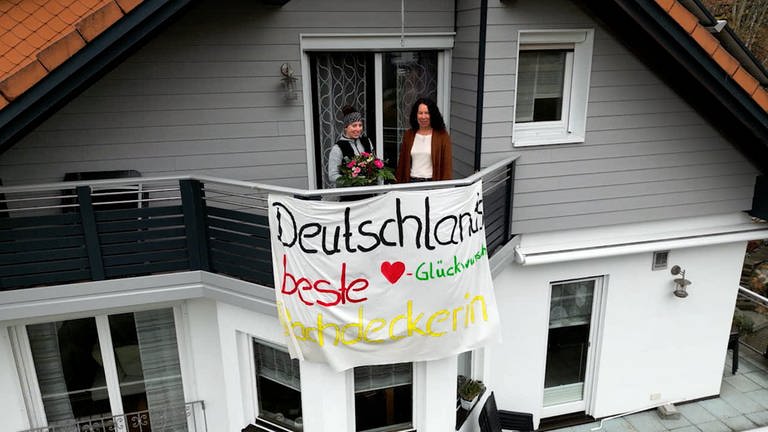 Jana Siedle steht mit einem Blumenstrauß neben ihrer Mama auf dem Balkon. Am Balkongeländer hängt ein großes Stoffbanner auf dem Deutschlands beste Dachdeckerin geschrieben steht. (Foto: SWR)