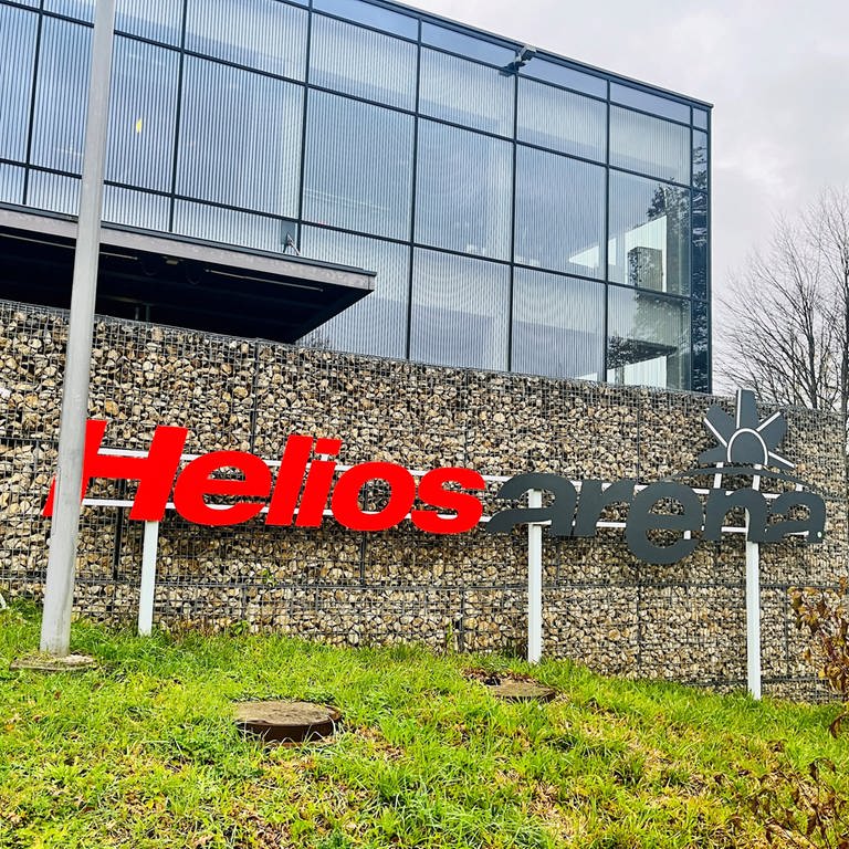 Vor einer Steinwand pragt groß das Logo der Helios-Arena, die große Eishalle in Villingen-Schwenningen, in der Spiele der Wild Wings stattfinden.  (Foto: SWR, Samantha Happ)