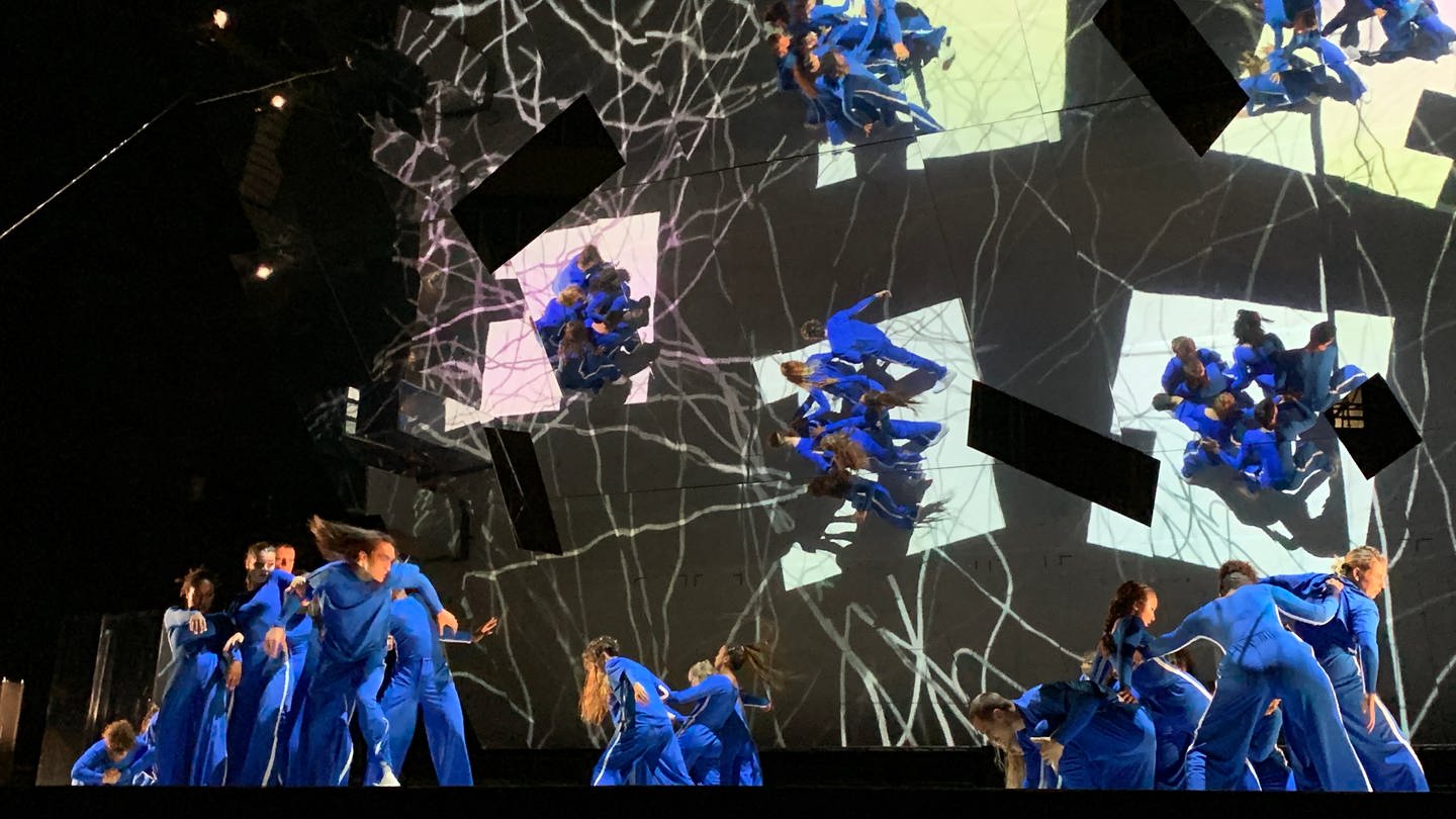 Tänzerinnen und Tänzer in blauen Anzügen stehen auf einer großen Bühne. Im Hintergrund ein überdimensionaler Spiegel. (Foto: SWR)