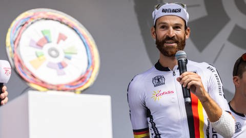 Simon Geschke steht im Vorfeld der dritten Etappe der Deutschland-Tour am Start in Freiburg und spricht in ein Mikrofon.