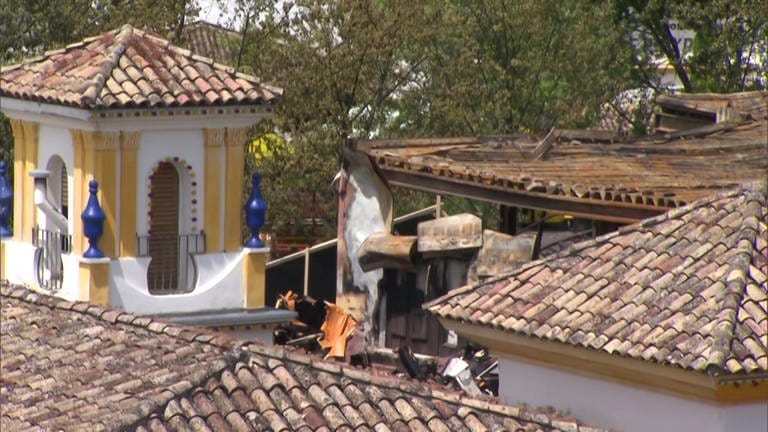 Zu sehen ist der Schaden nach dem Brand im Europapark. Das Bild ist von oben aufgenommen. Von einem Hausdach hängen angebrannte Metallstücke herunter. Links im Bild ist ein gelb-weißer Turm aus dem spanischen Themenbereich des Europaparks zu sehen.