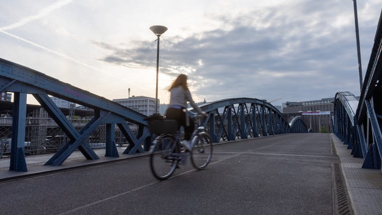 Eine Frau fährt auf einem Fahrrad über eine Freiburger Brücke, während im Hintergrund die Sonne aufgeht.