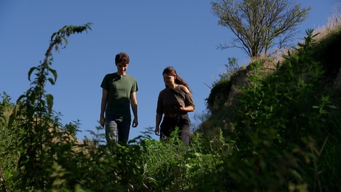 Ein junger Mann und eine junge Frau laufen vor blauem Himmel einen Feldweg hinab.