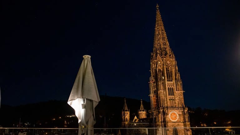 Das beleuchtete Freiburger Münster bei Nacht, im Vordergrund eine Terrasse mit eingeklappten Schirmen. (Foto: dpa Bildfunk, Philipp von Ditfurth)