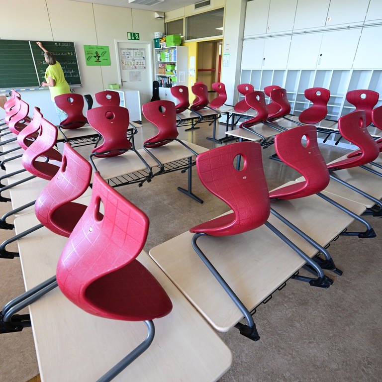 Stühle sind in einem Klassenzimmer einer Grundschule in Stuttgart auf Tische aufgereiht, im Hintergrund bereitet eine Lehrerin einen Tafelanschrieb vor. In der kommenden Woche ab dem 11. September beginnt in Baden-Württemberg nach den Sommerferien das neue Schuljahr.