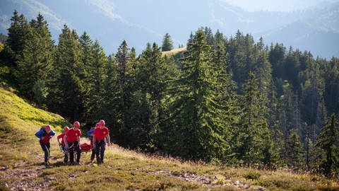 Mehrere Retter der Bergwacht sind im Einsatz. Sie tragen rote Helme und blaue Jacken. Im Hintergrund ist die malerische Kulisse des Schwarzwaldes zu sehen.