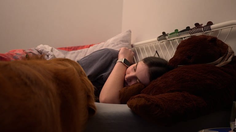 Eine Jugendliche liegt im Bett. daneben steht ein Golden Retriever und schaut sie an. (Foto: SWR)