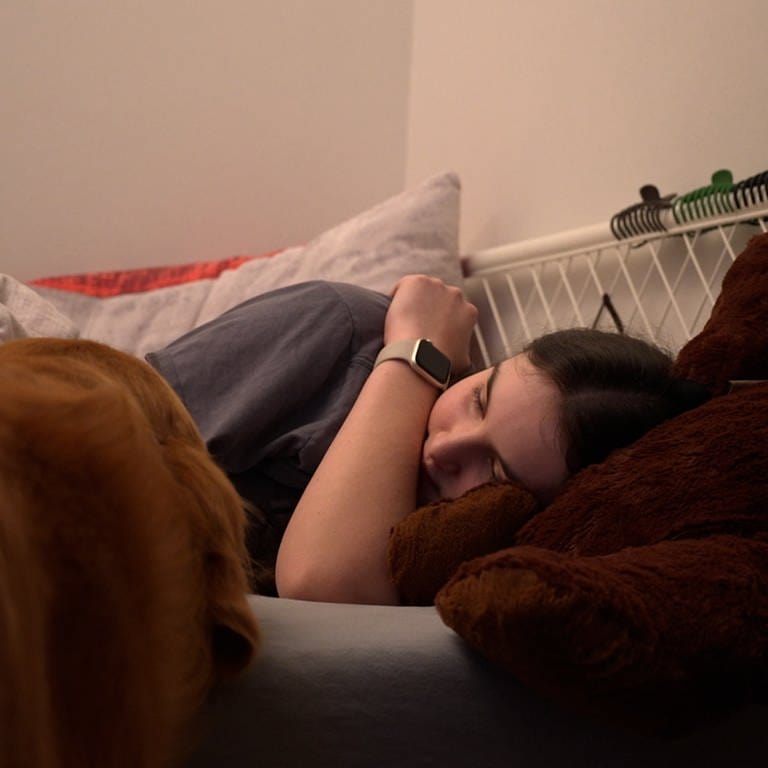 Eine Jugendliche liegt im Bett. daneben steht ein Golden Retriever und schaut sie an.