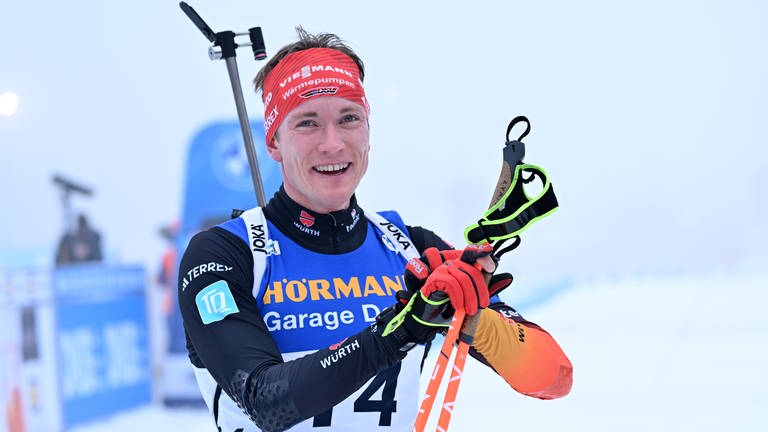 Mann mit Skistöcken und in Biathlon-Trikot schaut lachend in die Kamera. Er trägt ein rotes Stirnband mit Werbeslogan und auf seinem Rücken trägt er ein Biathlon-Gewähr.