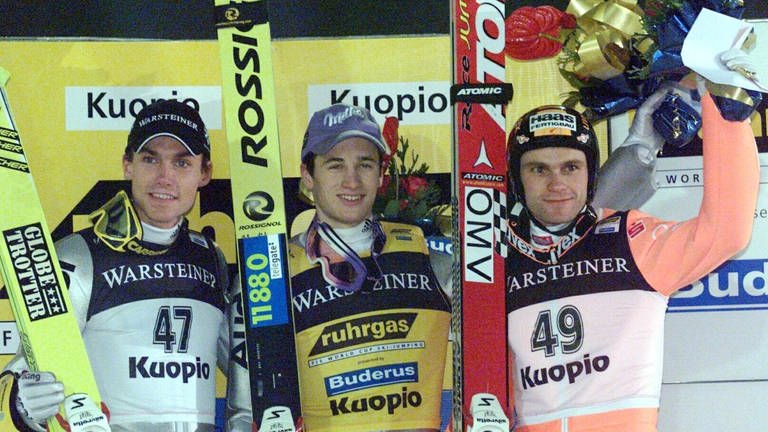 Drei Skispringer stehen mit ihren Skiern nebeneinander auf einem Siegerprotest. zwei halten große Blumensträuße hoch.