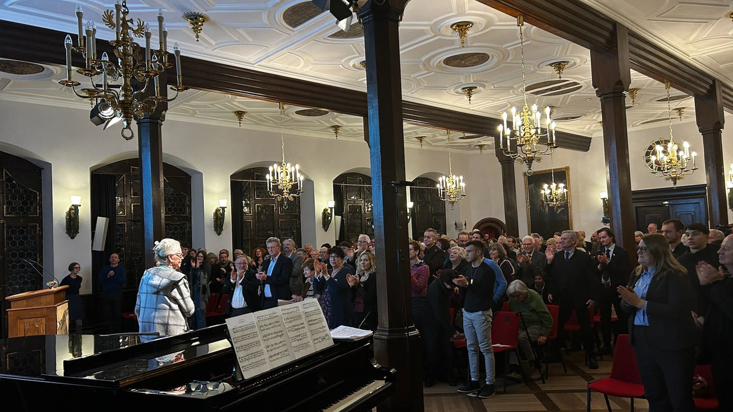 In einem großen Saal mit Säulen sitzen mehrere hundert Menschen. Vorne steht eine Frau mit weißen Haaren und dunkler Brille und hält einen Vortrag. (Foto: SWR)