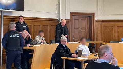 Im Gerichtssaal stehen alle Beteiligten neben ihren Sitzplätzen. Die Angeklagten Männer stehen ganz links. (Foto: SWR, Samantha Happ)