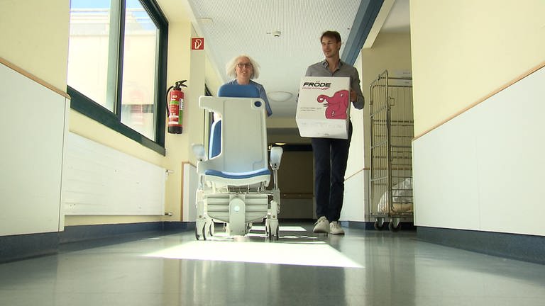 Zwei Mitarbeitende im Krankenhausflur beim Umzug, mit Kiste und medizinischem Stuhl