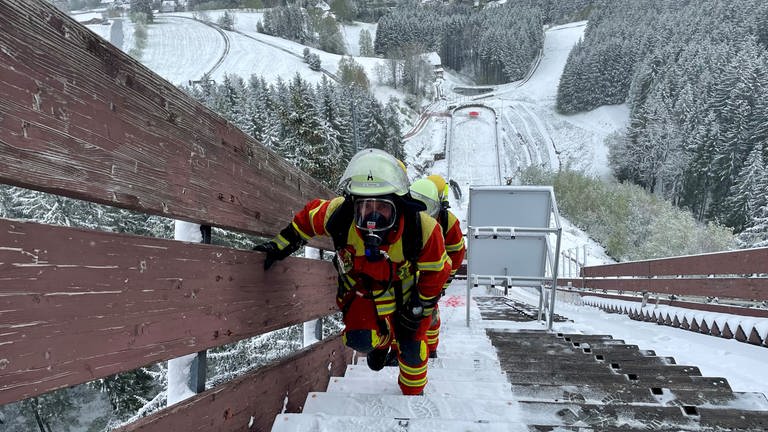 Zwei Feuerwehrleute laufen in voller Montur die verschneite Langenwaldschanze in Schonach hoch. (Foto: SWR)