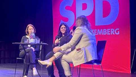 Drei Frauen sitzen vor SPD Leinwand auf einem Podium 