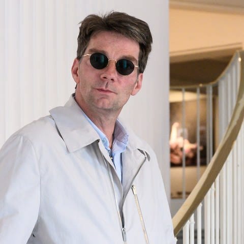 Mann mit runder Sonnenbrille und kurzen braunen Haaren schaut in die Kamera. Er trägt einen hellen Mantel und lehnt sich an ein Treppengeländer im Stil der 50er Jahre.