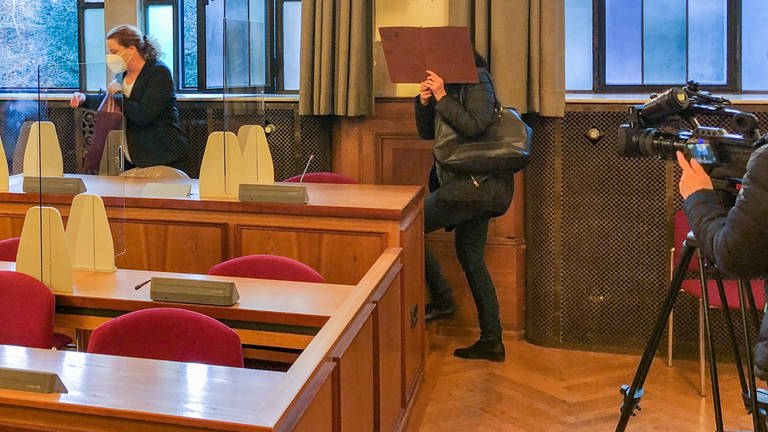 Eine Angeklagte verdeckt ihr Gesicht mit einer Pappe  einem Ordner, als sie das Landgericht Tübingen betritt. Sie soll ihr Kind getötet haben. Neben ihr ihre Anwältin, am Rand eine Videokamera eines Vertreters der Presse. (Foto: SWR, Stefanie Assenheimer)