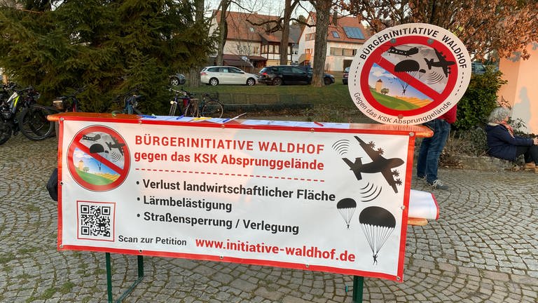Ein Plakat mit der Aufschrift  "Bürgerinitiative Waldhof gegen das KSK Absprunggelände" hängt quer vorne an einem Biertisch herunter.