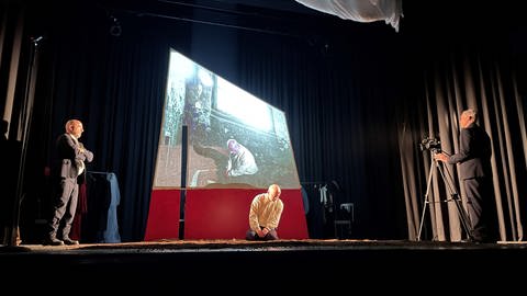 Auf eine halb durchsichtige Wand auf der Bühne werden historische Bilder und das aktuelle Bühnengeschehen projeziert. (Foto: SWR, Peter Binder)