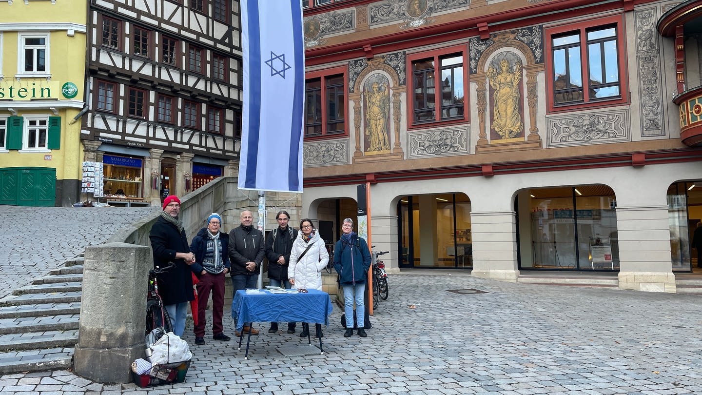 Sechs Menschen stehen vor einer gehissten Israel-Flagge. Im Hintergrund befindet sich das Tübinger Rathaus. Die sechs Mitglieder des Fördervereins für jüdische Kultur in Tübingen wollen dafür sorgen, dass die Israel-Flagge vor dem Rathaus diesmal hängen bleibt. (Foto: SWR, Theresa Krampfl)