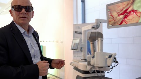 Holger Reinecke, Vorstandmitglied von Aesculap, zeigt ein Operationsmikroskop mit Roboterarm. (Foto: SWR, Mübeyra Erkus)