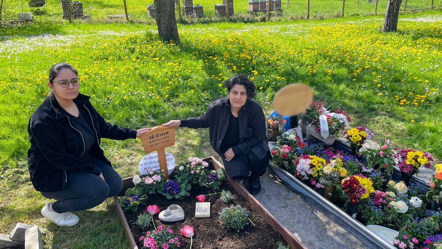 Am Grab ihres Mannes und Vaters knien links die 22-jährige Tochter und rechts die Mutter und halten die Hand auf das hölzerne Namensschild des Verstorbenen. Nebenan im Doppelgrab wurde die falsche Frau beerdigt. (Foto: SWR, Zundel, Mia)