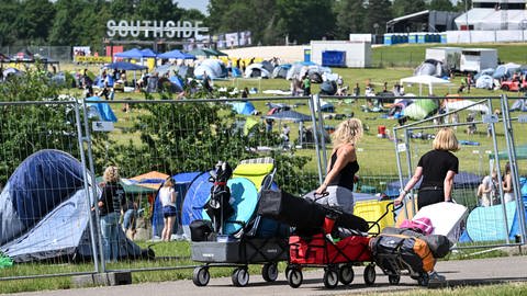 Zwei Festivalbesucherinnen ziehen mit Gepäck auf ihren Leiterwagen zum Festgelände, um einen Zeltplatz zu ergattern. 