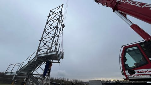 Das dreibeinige Grundgerüst des 18 Meter hohen Aussichtsturmes wird an Stahlketten per Autokran auf die betonierte Plattform gehievt (Foto: SWR, Richter, Tim)