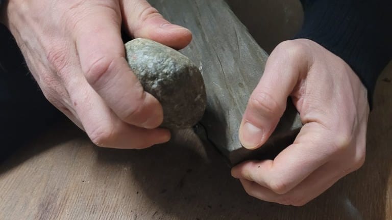 Man sieht zwei Hände. In beiden Händen befinden sich Steine, ein kleiner, ein großer. Mit dem kleinen Stein wird auf den großen geklopft. Es soll ein Steinwerkzeug entstehen. (Foto: Gregor Bader)