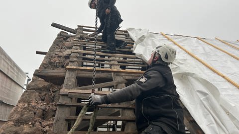 Am Dach tragen zwei Handwerker nacheinander Balken ab. Sie hängen sie an einen Kran. Danach werden die Balken nummeriert und für den Transport ins Christophstal vorbereitet. Wenn die Balken in schlechtem Zustand sind, werden sie repariert.