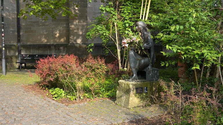 Man sieht eine Skulptur. Sie steht am Rand eines kleinen Weges in Tübingen. Man nennt sie: die "Sitzende". SIe hat einen Strauß Blumen im Arm. Die Blumen sind schon etwas verwelkt.
