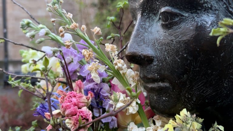 Man sieht den Kopf einer Statue. Links von ihr ist ein Blumenstrauß. Die Blumen sind schon leicht verwelkt. Bei der Statue handelt es sich um die "Sitzende" in Tübingen. 