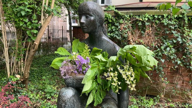 Man sieht eine Skulptur in Tübingen. Man nennt sie: die "Sitzende". Sie hat Blumen in den Arm gelegt bekommen. Viele davon sind grün. Man erkennt aber auch einen kleinen lila Strauß.