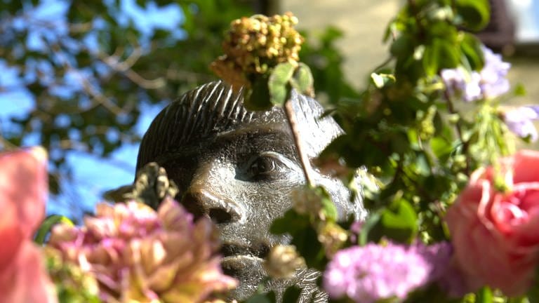 Man sieht den Kopf einer Skulptur. In Tübingen nennt man sie: die "Sitzende". Ihr werden oft Blumen in die Arme gelegt. Manchmal sieht man vor lauter Blümchen den Kopf gar nicht mehr. 