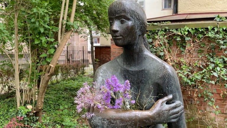 Man sieht eine Skulptur. In Tübingen nennt man sie: die "Sitzende". Ihr werden immer wieder Blumen in die Arme gelegt. Heute hat sie nur einen kleinen Strauss.
