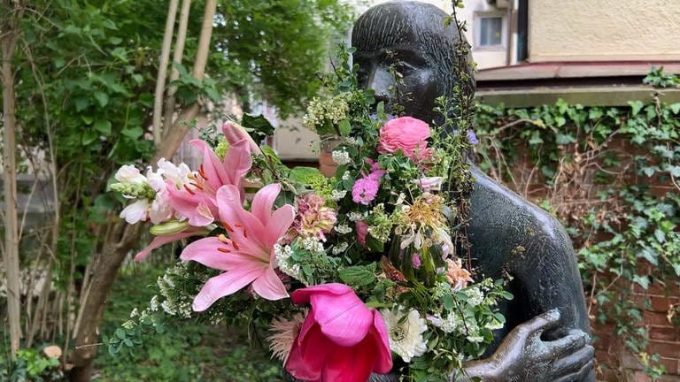 Man sieht eine Skulptur in einer Gasse in Tübingen. Sie hat einen großen Blumenstrauß in den Armen. Man nennt sie die "Sitzende". In Tübingen wundert man sich: Wer kümmert sich so liebevoll um die "Sitzende" und schenkt ihr Blumen? 