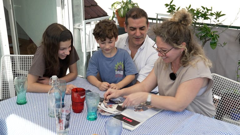 Die Familie Cardosa Zea  Markovic sitzt an einem Tisch auf dem Balkon und schaut Erinnerungsfotos an. Die Eltern haben sich bei einem Erasmus Semester kennengelernt