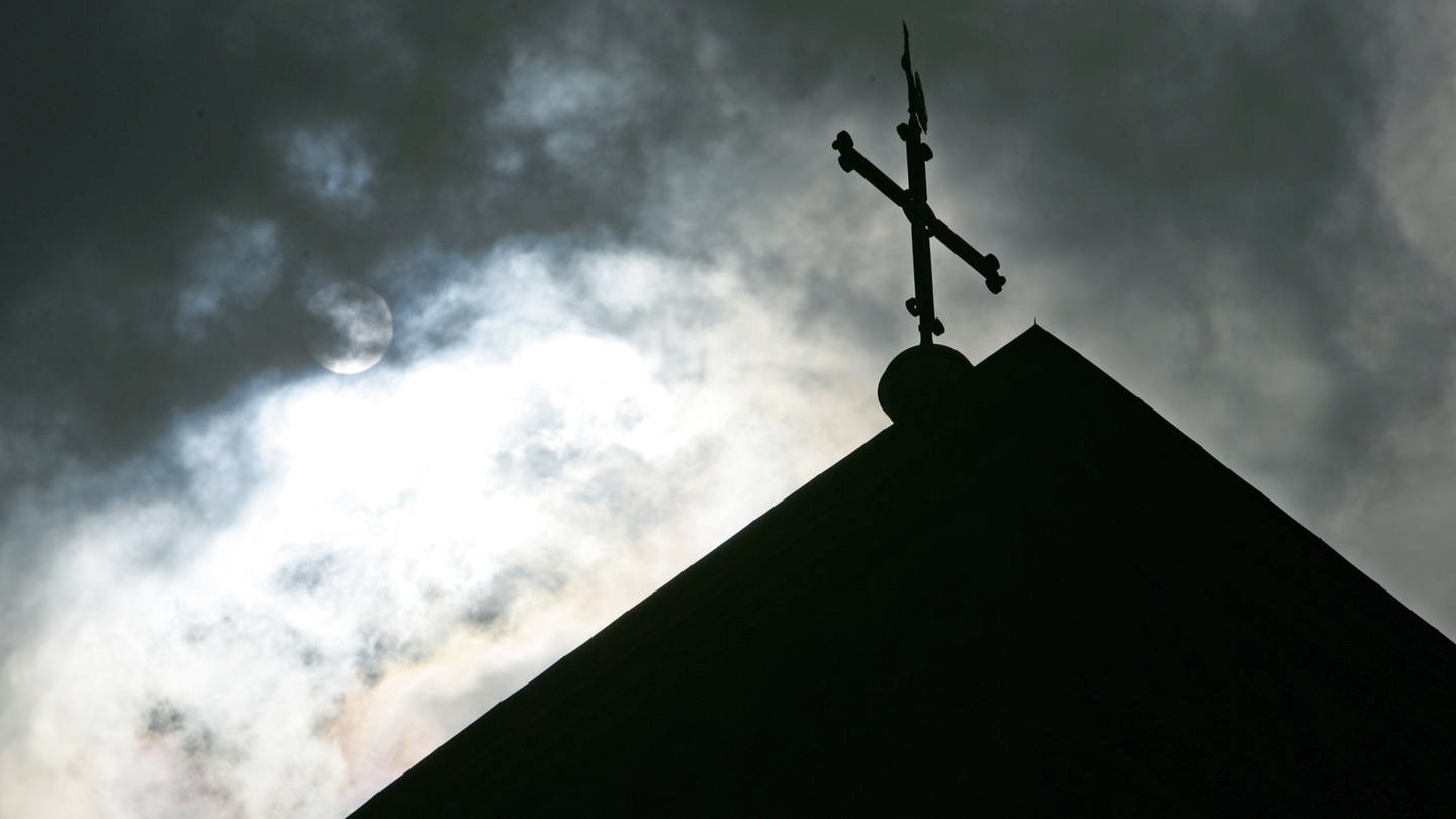 Im Gegenlicht und vor wolkenverhangenem Himmel ist eine Kirchturmspitze mit Kreuz zu sehen. (Foto: dpa Bildfunk, picture alliance/dpa | Friso Gentsch)