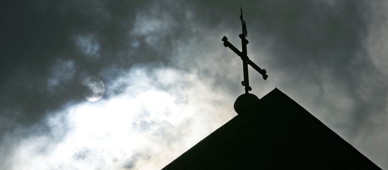 Im Gegenlicht und vor wolkenverhangenem Himmel ist eine Kirchturmspitze mit Kreuz zu sehen. (Foto: dpa Bildfunk, picture alliance/dpa | Friso Gentsch)